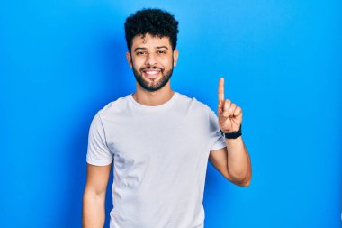 Sakallı, günlük beyaz tişörtlü genç bir Arap bir numaralı parmağını gösterip gösterirken kendinden emin ve mutlu bir şekilde gülümsüyor.. 