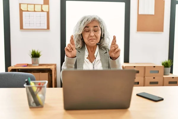 中年女商人坐在写字台上 用笔记本电脑在办公室里摆弄着手指 带着希望微笑 闭上眼睛 幸运与迷信的概念 — 图库照片