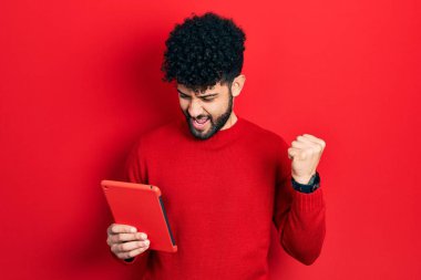 Sakallı genç Arap adam dokunmatik bir cihaz kullanıyor. Gururla bağırıyor, zaferi kutluyor ve başarıyı kollarını kaldırarak kutluyor. 