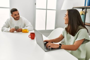 Laptop ve akıllı telefon kullanan genç latin çift evde kahve içiyor..