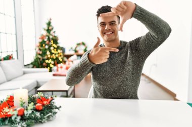 Genç İspanyol adam Noel ağacının yanında masada oturuyor gülümsüyor ve elleri ve parmakları mutlu yüzle çerçeve yapıyor. yaratıcılık ve fotoğrafçılık kavramı. 