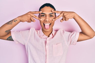 Genç Afrikalı Amerikalı adam zafer işareti yapıyor ve barış jesti komik bir ifadeyle dilini dışarı çıkarıyor.. 