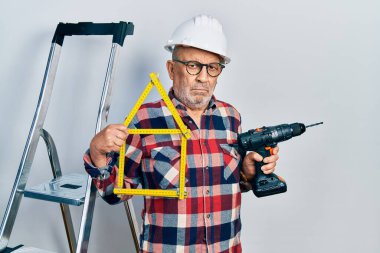 Elinde tornavida tutan yakışıklı bir adam inşaat merdivenlerinin yanında şapka takıyor. Şüpheci ve gergin. Problemden dolayı surat asıyor. negatif kişi. 