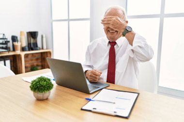 Ofiste çalışan kıdemli bir adam bilgisayar laptopu kullanıyor. Gülümsüyor ve elleri yüzün üzerinde gülüyor. Şaşırtmak için gözleri örtüyor. kör kavram. 