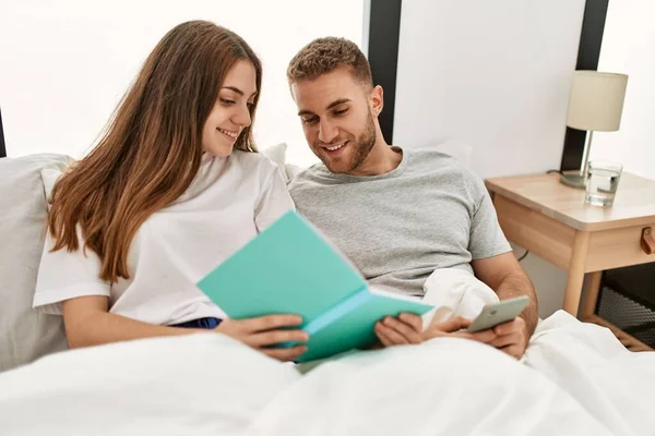 Unga Kaukasiska Par Läsa Bok Och Använda Smartphone Sängen — Stockfoto