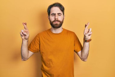 Sakallı, sarı tişörtlü beyaz bir adam. Umut dolu ve gözleri kapalı bir şekilde parmaklarıyla işaret ediyor. Şans ve batıl inançlar. 