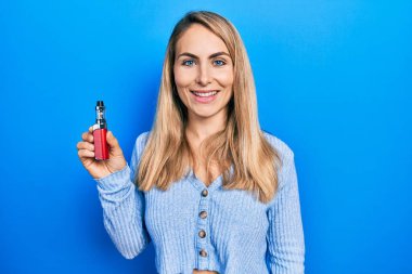 Elektronik sigara tutan genç beyaz kadın pozitif ve mutlu görünüyor ve gülümsüyor dişlerini gösteren bir gülümsemeyle. 