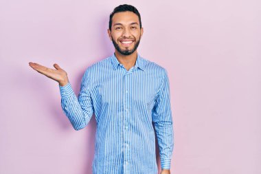 Sakallı, sıradan mavi tişörtlü, neşeli gülümseyen, elini kaldırıp kameraya bakan İspanyol bir adam.. 