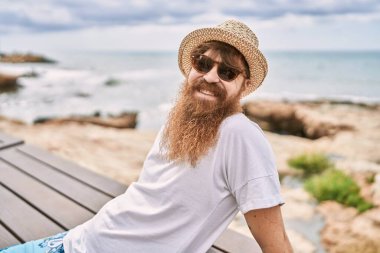 Genç, kızıl saçlı, gülümseyen turist kumsaldaki bankta mutlu bir şekilde oturuyor..