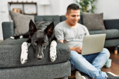 Genç İspanyol adam evde köpekle yerde otururken dizüstü bilgisayar kullanıyor..
