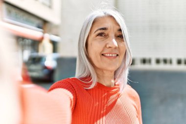 Orta yaşlı, gri saçlı, gülümseyen bir kadın şehrin kamerasının önünde selfie çekerken mutlu oluyor..