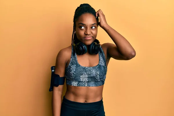 African american woman with braided hair wearing sportswear and arm band confuse and wonder about question. uncertain with doubt, thinking with hand on head. pensive concept.