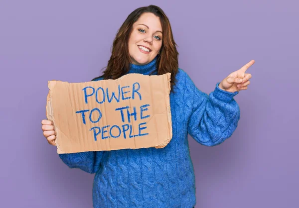 Young plus size woman holding power to the people banner smiling happy pointing with hand and finger to the side