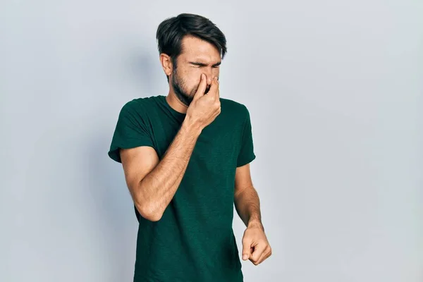 若いヒスパニック系の男性は 鼻の上に指で息を保持し 臭いと嫌な 耐えられない臭い何かを嗅ぐカジュアルな白いTシャツを着て 臭いが悪い — ストック写真