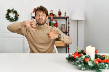 Noel süslemesi masasında oturan sakallı yakışıklı genç adam baş parmak kaldırıyor, anlaşmazlık ve anlaşma ifadesi takınıyor. çılgın çatışma 