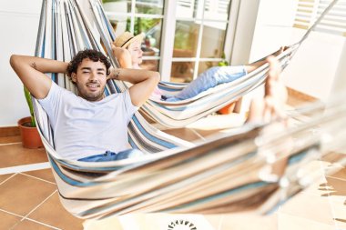 Mutlu gülümseyen genç çift, elleri başlarının üzerinde terasta uzanmış bir şekilde rahatlar..