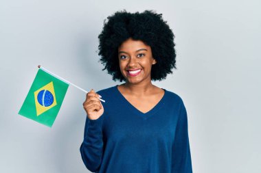 Brezilya bayrağı tutan genç Afro-Amerikalı kadın pozitif ve mutlu görünüyor ve kendine güvenen bir gülümsemeyle dişlerini gösteriyor. 