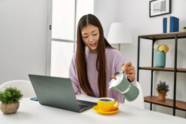 Genç Çinli kız dizüstü bilgisayar kullanıyor ve kahve içiyor. Evdeki masada oturuyor..