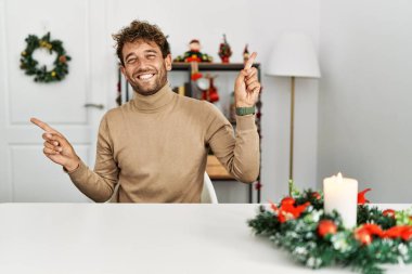 Noel süslemesi masasında oturan sakallı, kendine güvenen, farklı yönleri gösteren genç ve yakışıklı bir adam. reklam için kopyalama alanı 