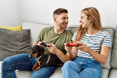 Genç İspanyol çift gülümseyerek evde köpekle kanepede oturup video oyunu oynuyor..