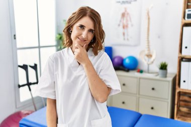 Orta yaşlı fizyoterapist kadın ağrı kesici kliniğinde çalışıyor kameraya kendinden emin bakıyor kollarını kavuşturmuş ve çenesini kaldırmış bir şekilde gülümsüyor. Olumlu düşünmek. 