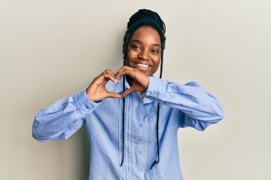 Saçları örgülü, mavi gömlekli, aşık gülümseyen, elleriyle kalp sembolü yapan Afro-Amerikalı bir kadın. romantik kavram. 