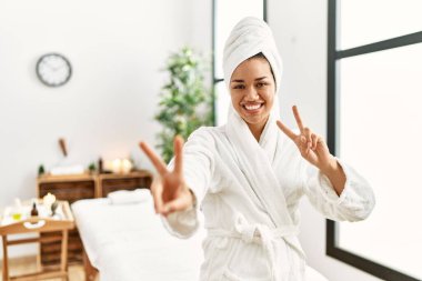 Havlu ve bornoz giymiş genç esmer kadın güzellik merkezinde durup kameraya bakıp zafer işareti yapıyor. İki numara.. 