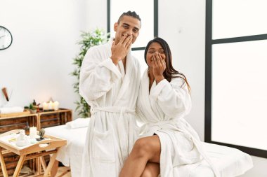 Young latin couple wearing towel standing at beauty center laughing and embarrassed giggle covering mouth with hands, gossip and scandal concept 