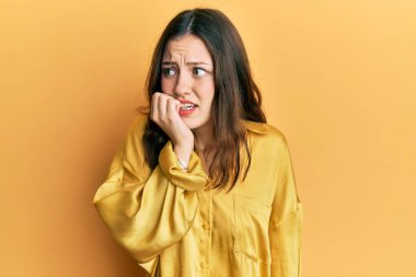 Genç esmer kadın gündelik sarı tişört giyiyor. Stresli ve gergin görünüyor. Elleri ağızdan tırnak yiyor. anksiyete. 