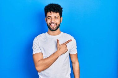 Sakallı, beyaz tişörtlü, neşeli, yüzünde bir gülümseme olan, mutlu ve doğal bir ifadeyle elini ve parmağını kaldırmış genç bir Arap. 