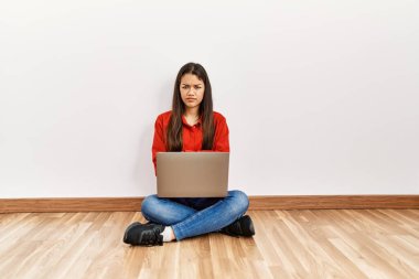 Genç esmer kadın boş bir odada dizüstü bilgisayarlı şüpheli ve gergin bir şekilde oturuyor. Yüzünde çapraz kollu tasvip etmeyen bir ifade var. negatif kişi. 