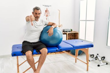 Orta yaşlı İspanyol bir adam, ağrı kesici kliniğinde elinde pilates topu tutarken mutsuz ve sinirli görünüyor. Reddedilmiş ve olumsuz jestler yapıyor. kötü ifade. 