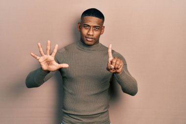 Balıkçı yaka kazak giyen genç siyahi bir adam altı numaralı parmaklarını gösterip onu gösterirken kendinden emin ve mutlu bir şekilde gülümsüyor.. 