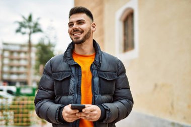 Yakışıklı, sakallı, mutlu ve kendine güvenen, akıllı telefon kullanan, kışlık palto giyen bir İspanyol.