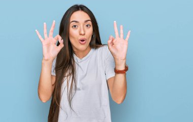Gündelik beyaz tişört giyen genç İspanyol kız şaşırmış ve şok olmuş bir şekilde parmaklarıyla onay sembolü yapıyor. çılgın ifade 