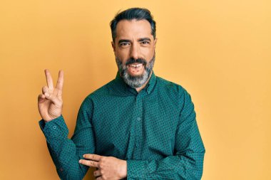 Sakallı, gri saçlı, iş kıyafetleri giyen, kameraya gülümseyen ve zafer işareti yapan orta yaşlı bir adam. İki numara.. 