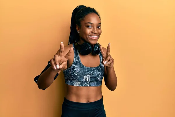 African american woman with braided hair wearing sportswear and arm band smiling looking to the camera showing fingers doing victory sign. number two.