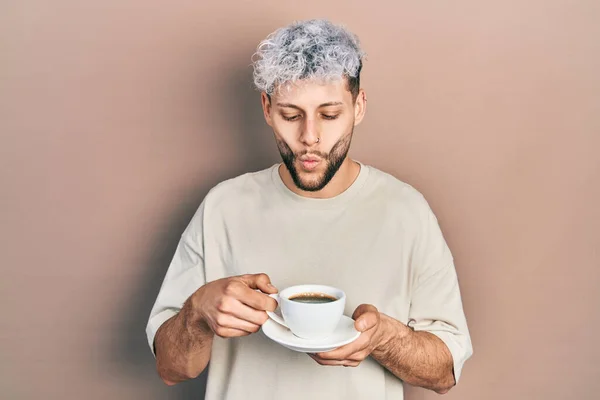 Young hispanic man with modern dyed hair drinking a cup of coffee making fish face with mouth and squinting eyes, crazy and comical.
