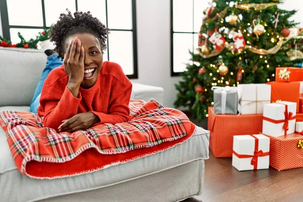 若いアフリカ系アメリカ人の女性によってソファの上に横たわっていますクリスマスツリーで手で一つの目を覆います 顔に自信を持って笑顔と驚きの感情 — ストック写真