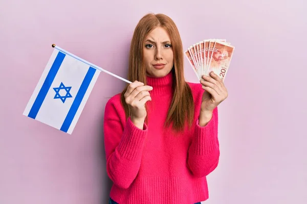 年轻的爱尔兰女人拿着以色列国旗和谢克尔钞票 表示怀疑和紧张 因为问题皱着眉头 消极的人 — 图库照片