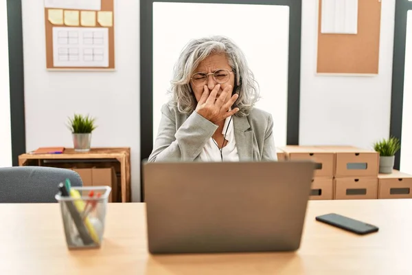 Middle age businesswoman sitting on desk working using laptop at office smelling something stinky and disgusting, intolerable smell, holding breath with fingers on nose. bad smell
