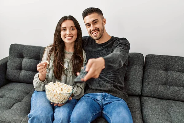 Young hispanic couple watching film and eating popcorn sitting on the sofa at home.