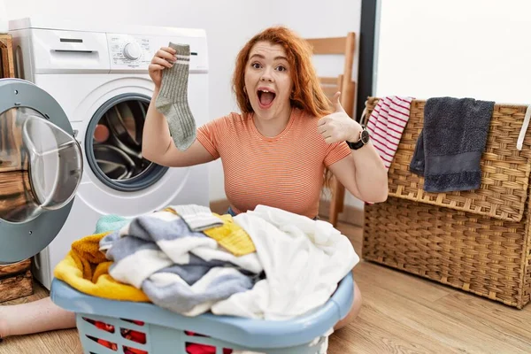 若い赤毛の女性は手で幸せな親指を行う洗濯機に汚れた洗濯機を入れてジェスチャー 成功を示すカメラを見ると — ストック写真