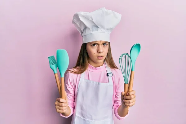 美しいブルネットの女の子は 問題のために動揺を眉をひそめて 懐疑的で緊張した調理器具を保持するプロのコックエプロンを身に着けています 否定的な人 — ストック写真