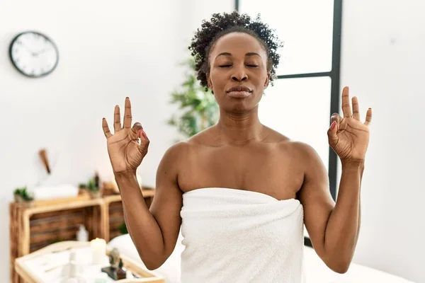 美容センターに立つタオルを身に着けている若いアフリカ系アメリカ人女性は 指で瞑想ジェスチャーを行う目を閉じてリラックスして笑顔 ヨガのコンセプト — ストック写真