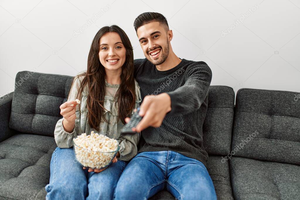 Young hispanic couple watching film and eating popcorn sitting on the sofa at home.