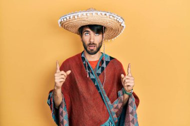 Meksika şapkası tutan genç İspanyol adam şaşırmış ve şaşırmış bir şekilde yukarı bakıyor ve parmaklarıyla işaret ediyor ve kollarını kaldırıyor.. 