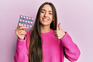 Genç İspanyol kız elinde haplarla mutlu ve pozitif gülümsüyor, başparmağıyla mükemmel ve onay işareti yapıyor. 