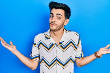 Sıradan kıyafetler giyen, elleri ve kolları havada, kafası karışmış genç İspanyol bir adam. şüphe kavramı. 