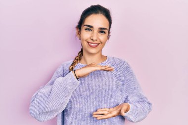 Latin kökenli genç bir kız, sıradan kıyafetler giyiyor. Elleriyle işaret ediyor. Ölçü sembolü. Kameraya bakıp gülümsüyor. ölçüm kavramı. 